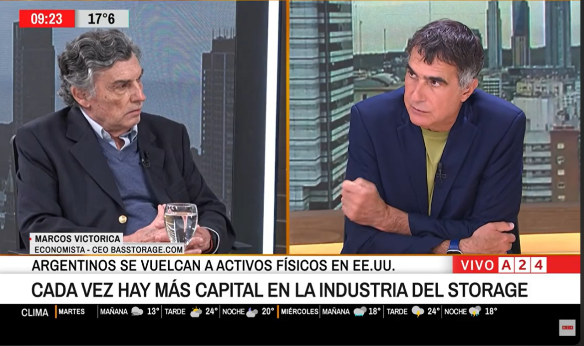 Split-screen TV interview: on the left, an older man in a blazer; on the right, a man in a blue blazer with a lime-green shirt; a newsroom backdrop; a ticker at the bottom about the storage industry.