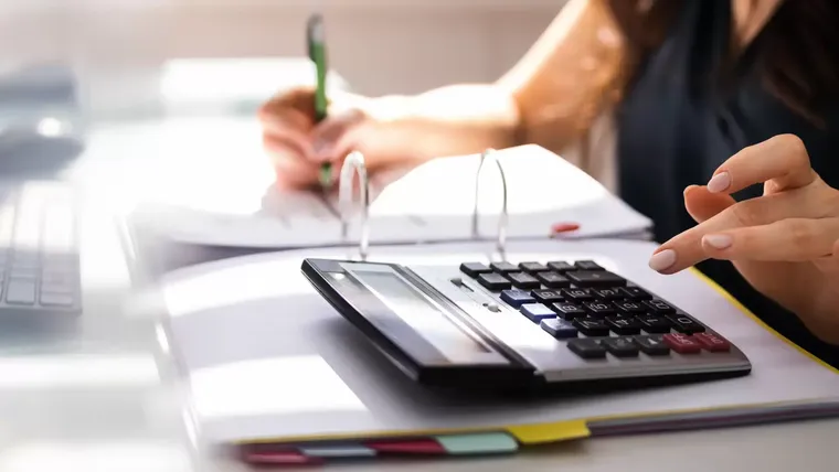Person uses a calculator on a desk while writing in a binder with a pencil; business/office work context.