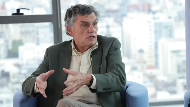 Man in a green blazer speaks and gestures during an interview in a bright office with a city view.